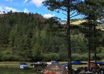 Vehicles and tents are scattered across a grassy field backed by tree covered hills.