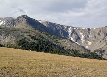 A photo of a meadow with dry brown grass in the foreground and a mountain in the background with patches of snow, partly cloudy sky.