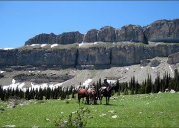 Steep limestone cliffs tower over alpine meadows and scattered spruce trees. A man in a cowboy hat with three horses sits on a horse in the foreground.