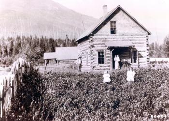 Historic black and white photo showing family standing in front of two story log cabin with mountain in the distance