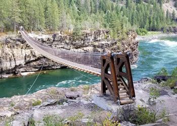 Suspension bridge over a flowing river with rapids with forested mountains in background