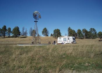 A lone, small RV camps next to a windmill in tall grass with scattered ponderosa pine trees in the background.