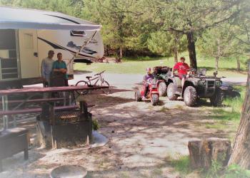 A couple stands in the shade next to their RV while kids sit on ATVs at their campsite.