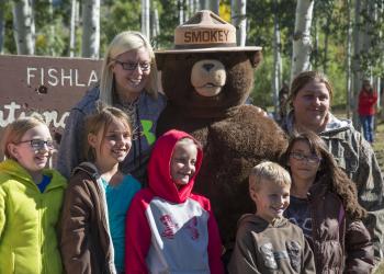 A photo of 5 kids and two adults standing next to smokey bear in the outdoors. A Fishlake National Forest sign is behind them.