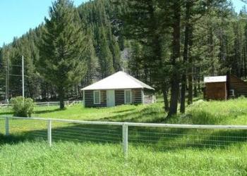 A brown log cabin with white door sitting on a grassy plain behind a fence