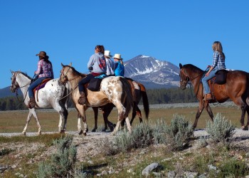 Five people on horses, including two spotted appaloosas, ride along a prairie with steep rocky mountains in the background.
