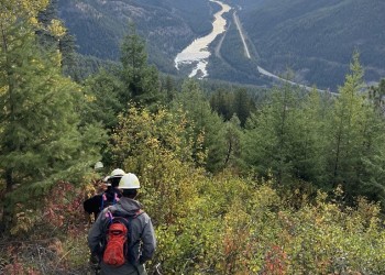 3 people walk through mountain side covered in trees and shrubs with river, highway and train tracks in background.