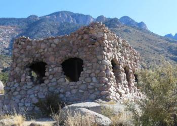 A shelter made of rocks with holes in the walls for windows, nestled beneath rocky and forested mountain peaks.