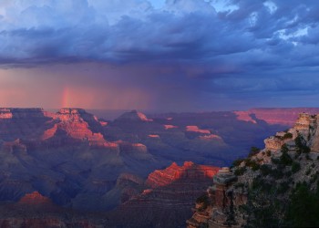 Sunset view of the Grand Canyon with storm clouds overhead and sheets of rain and a partial rainbow in the distance.
