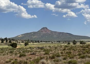 A lone rocky peak rises above a grassy plain beneath a partially cloudy sky.