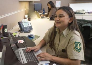 two Forest Service employees in uniform at a desk on computer