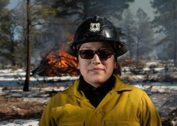 woman in Nomex fire shirt with hard hat standing in front of burning pile