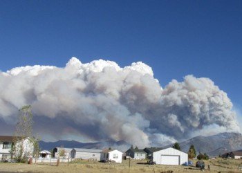 Large plume of smoke from a wildfire hangs over residential homes in a rural landscape.