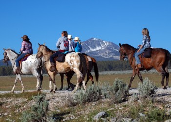 Five people on horses, including two spotted appaloosas, ride along a prairie with steep rocky mountains in the background.