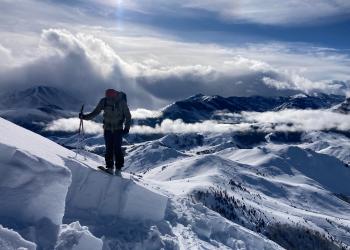 A skier stands on the edge of a wall of stable snow where an avalanche was triggered. Behind the skier, lower peaks covered with snow intertwine with clouds. Above the clouds, the sun shines brightly in the blue sky, casting shadows on the right faces of the mountains below.