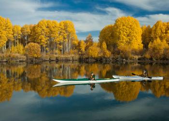 Two people kayak through the Deschutes National Forest and fall foliage is reflected on the water