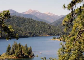 A lake is framed by trees on all sides. Mountains are in the background.