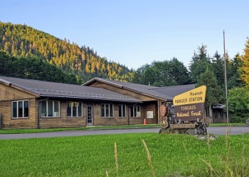 green grass in the foreground, brown building with a FS sign yellow and brown saying Hoonah Ranger District with green evergreen trees and mountain in background